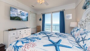 A bedroom with a bed featuring a sea-themed bedspread, a ceiling fan, a television on the wall, and a view of the ocean through the window. - Moon Tide|Oceanfront Showplace|Dog Friendly|Elevator|Rooftop Deck|Family-Friendly Amenities (Edisto Beach)