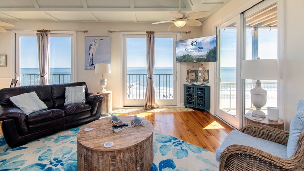A living room with a leather sofa, a round wooden coffee table, and a wicker chair. There are large windows with curtains, a ceiling fan, and a view of the ocean. - Moon Tide|Oceanfront Showplace|Dog Friendly|Elevator|Rooftop Deck|Family-Friendly Amenities (Edisto Beach)
