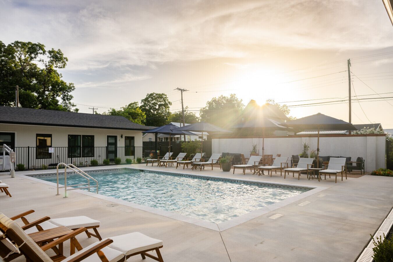 A swimming pool with lounge chairs and umbrellas, surrounded by a building with a white exterior and a black roof.