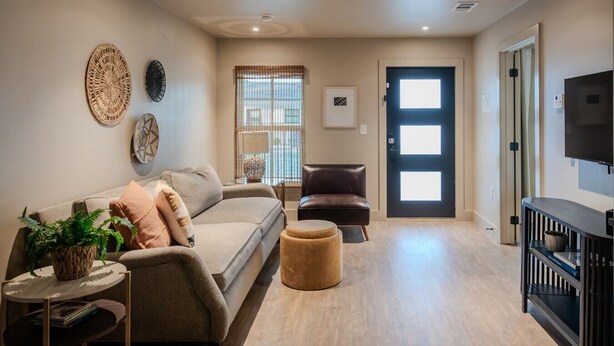 A modern living room with a sofa, a coffee table, a side table, a small round ottoman, a TV on a stand, and a door with a window. - Gloria Inn 2 -5 minutes from Baylor and 2 Blocks to Magnolia (Waco)