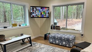 Una habitación acogedora con un amplio ventanal, un televisor de pantalla plana, una mesita, un otoman con estampado y una silla.