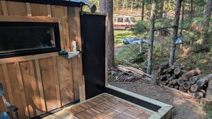 Una terraza de madera con una puerta negra, una silla y un cobertizo pequeño en un área boscosa.