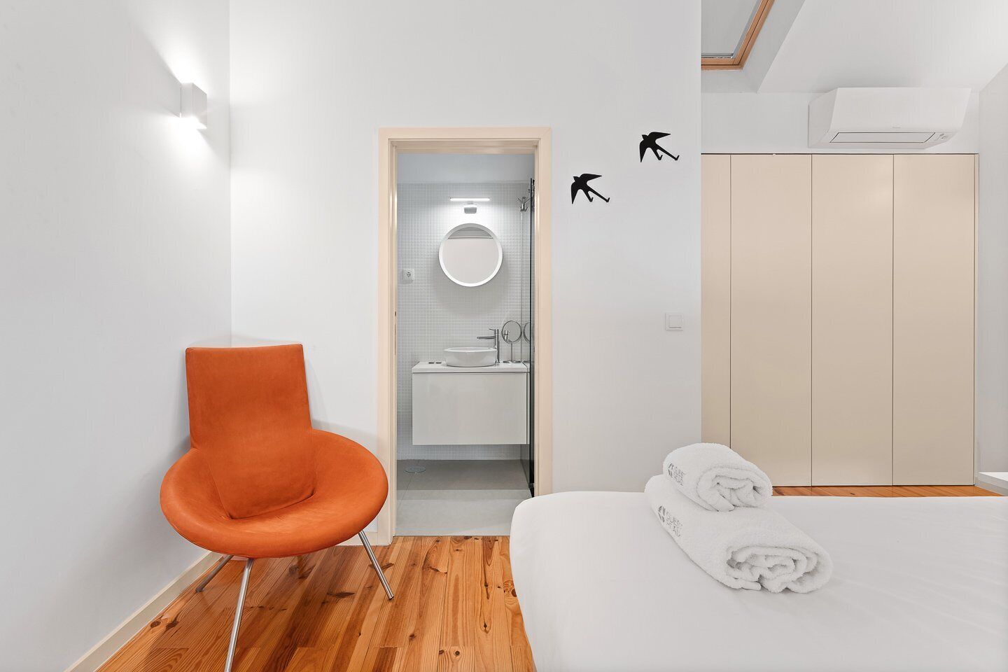 A modern bedroom with a wooden floor, a white bed, an orange chair, a wardrobe, and a bathroom visible through an open door.