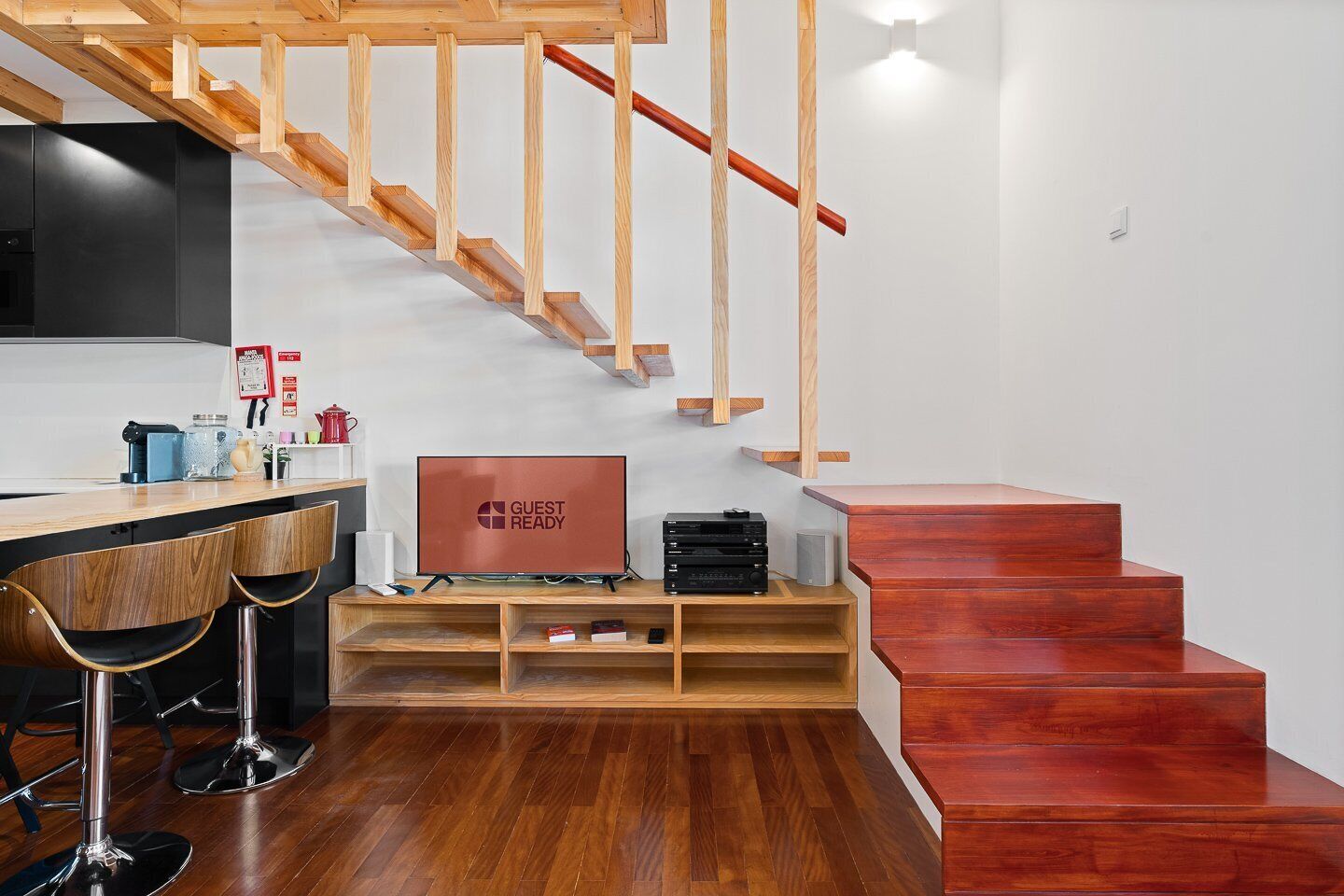 A modern kitchen with wooden flooring, a built-in wooden staircase, and a flat-screen TV on a wooden stand.