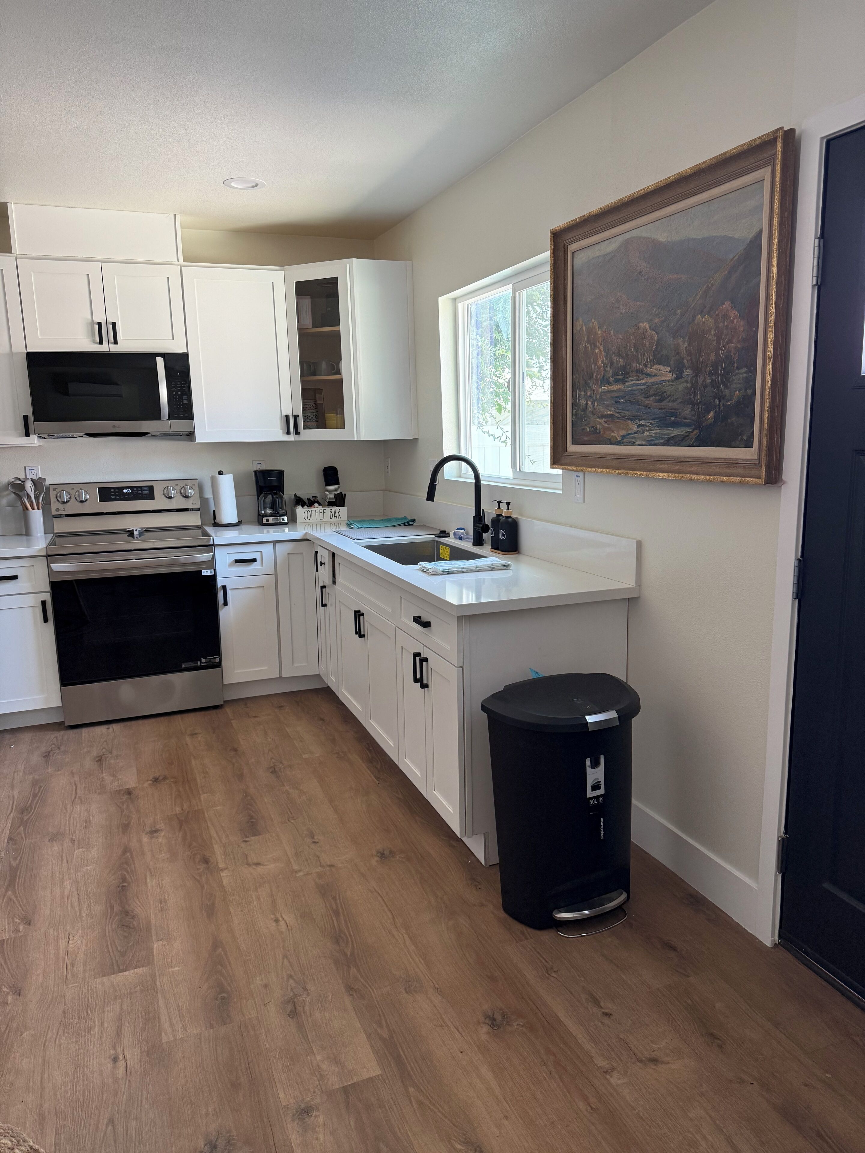 A kitchen with white cabinets, stainless steel appliances, a black trash bin, and a painting on the wall.