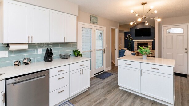 A modern kitchen with white cabinets, a dishwasher, and a built-in oven. There is a dining area with a table and chairs, and a living room with a fireplace. - Modern Comfort in Des Moines` West Side 4BR/2.5BR (Clive)