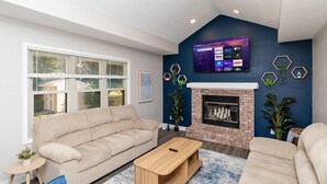 A living room with a beige sofa, a wooden coffee table, a blue rug, a fireplace, and a TV mounted on the wall displaying various streaming services. - Modern Comfort in Des Moines` West Side 4BR/2.5BR (Clive)