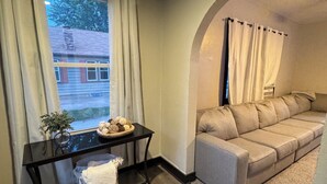 Un salon chaleureux avec un canapé, une table basse agrémentée d’une plante, et une vue sur une maison par la fenêtre.