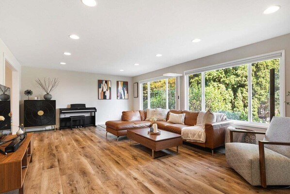 A spacious living room with a large sectional sofa, a coffee table, and a piano. - Perch - Spacious 4-Bed Home w/ Office, Gym & Yard (Redmond)