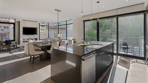 A modern kitchen with a large island, dining area, and large windows offering a view of the outdoors.