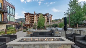 A fire pit area with seating, surrounded by stone walls, in front of a multi-story building with balconies.