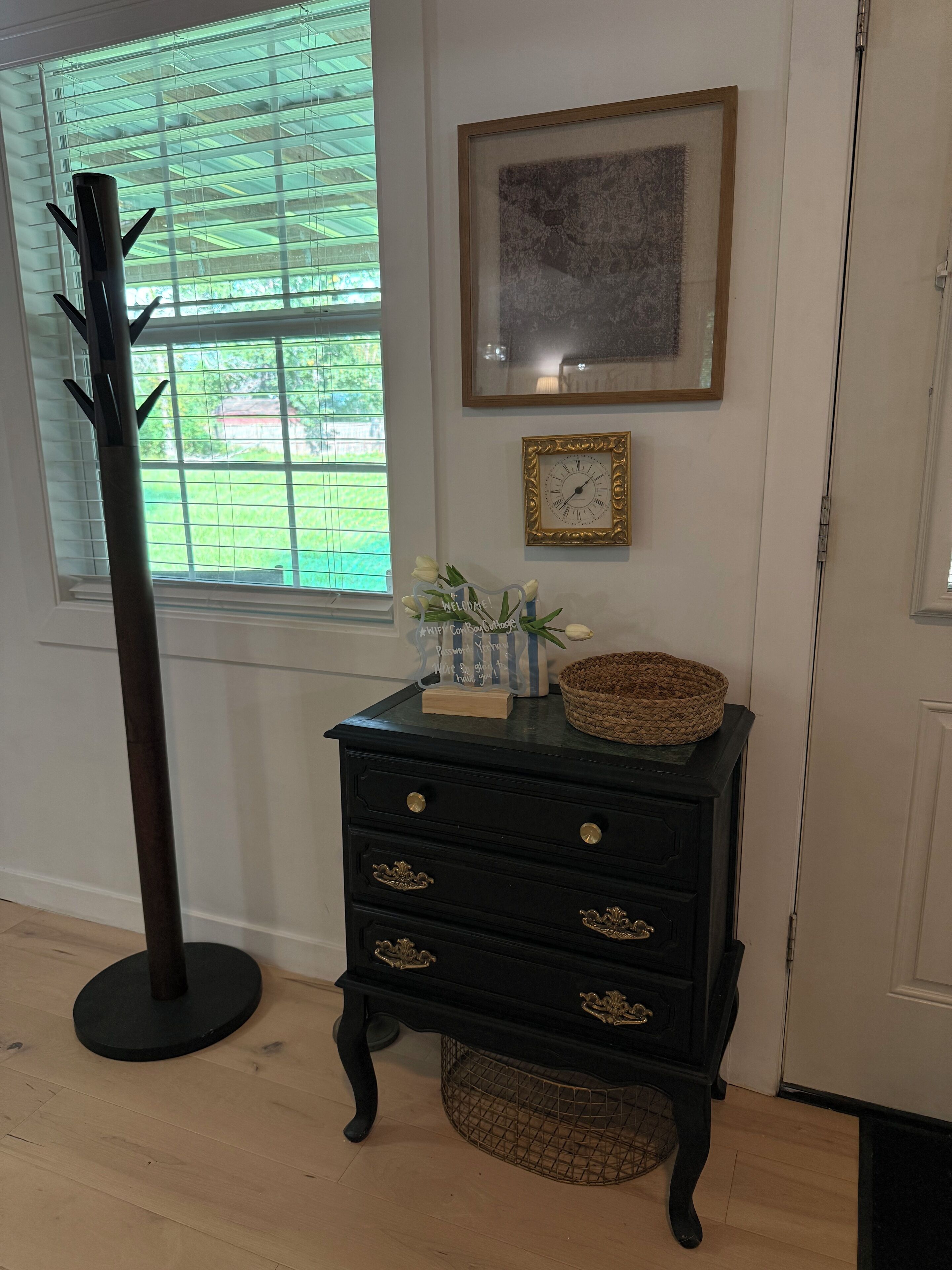 Une pièce avec une commode noire, une horloge, un tableau encadré et un grand lampadaire.