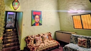 A room with a brick ceiling, a sofa with patterned cushions, a painting on the wall, and a window with blinds.