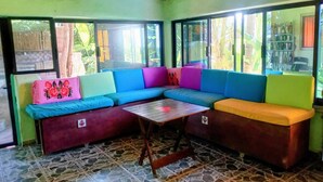 A room with a colorful sectional sofa, a small wooden table, and large windows with a view of greenery outside.