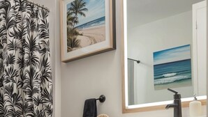 A bathroom with a toilet, a shower curtain with a leaf pattern, wooden cabinets, a mirror, and framed beach paintings on the wall.