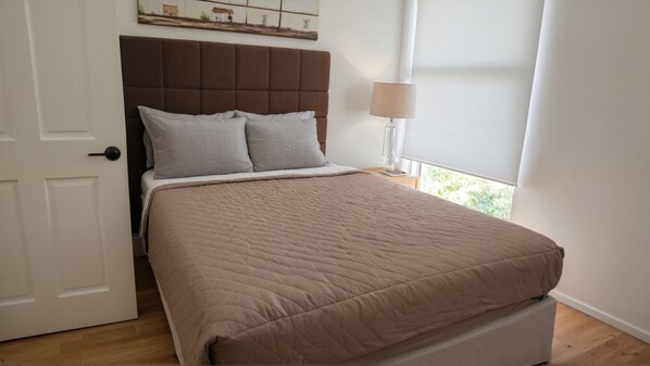 A bedroom with a bed, a headboard, a nightstand, a lamp, and a window with blinds.