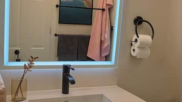 A bathroom with a white sink, a mirror with a light, a towel rack, and a toilet paper holder.