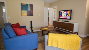 A living room with a blue sofa, red and yellow pillows, a yellow throw blanket, a TV on a wooden cabinet, and a painting on the wall.