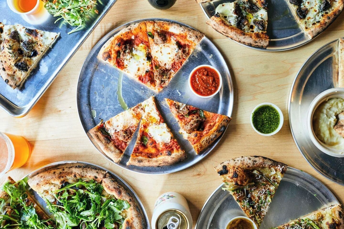 Un assortiment de pizzas servies sur des plateaux en métal, accompagnées de différentes sauces et d’une boisson.
