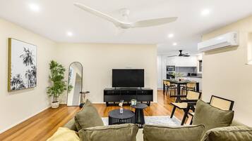 A modern living room with a sofa, a television, a dining area, and a kitchen in the background.