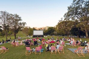 Un espace extérieur aménagé pour la projection de films, avec un grand écran, des rangées de chaises rayées et un champ d’herbe.