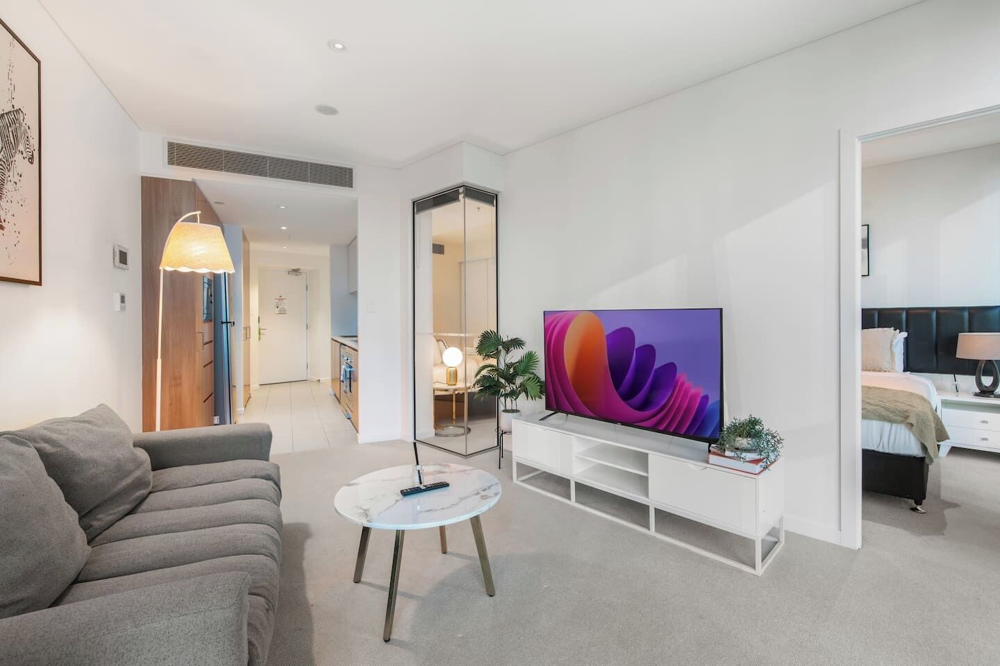 A modern living room with a flat-screen TV, a grey sofa, a coffee table, and a hallway leading to a bedroom.