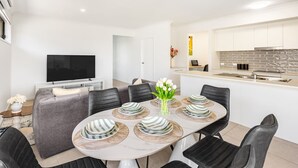 Een moderne eetruimte met een ronde tafel gedekt voor zes personen, omringd door zwarte stoelen. Een flatscreen televisie is links op de muur bevestigd.