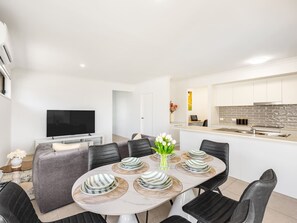 A modern dining area with a round table set for six, surrounded by black chairs. A flat-screen TV is mounted on the wall to the left.