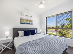 A modern bedroom with a large bed, a nightstand, a wall-mounted air conditioner, and a window with a view of the outside.