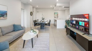 A modern living room with a sofa, coffee table, TV, and a dining area with chairs and a table. - Modern 2BR Apartment Balcony & Central Convenience (Macgregor)