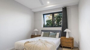 A bedroom with a bed, two bedside tables, a window with curtains, and a view of trees outside. - Modern 2BR Apartment Balcony & Central Convenience (Macgregor)