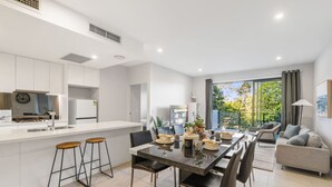 A modern kitchen with white cabinets, a dining area with a dark table, and a living room with a sofa and a balcony with a view. - Modern 2BR Apartment Balcony & Central Convenience (Macgregor)