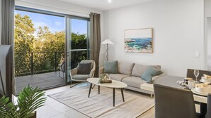 A modern living room with a sofa, armchairs, a coffee table, and a dining table set for two. A large window offers a view of trees and a balcony. - Modern 2BR Apartment Balcony & Central Convenience (Macgregor)