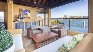 Un área de estar al aire libre techada con mobiliario de mimbre, una mesa de centro de madera y vistas al agua y edificios al fondo.