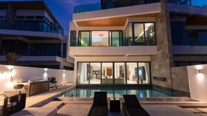 A modern multi-story building with a swimming pool, outdoor seating, and a well-lit interior. - Beachfront Villa 3BR Infinity Pool V38 (Rawai)