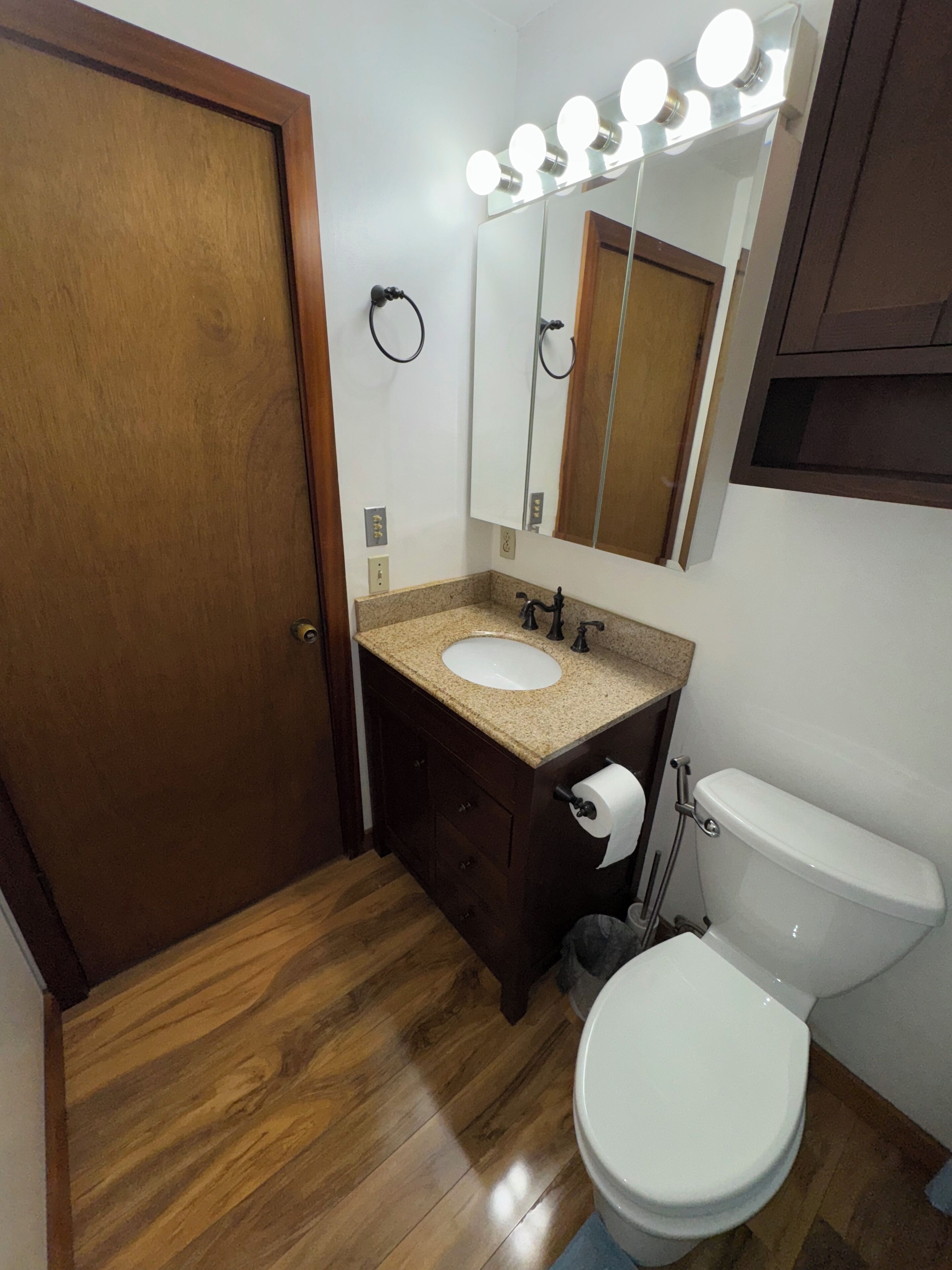 A bathroom with a wooden door, a sink with a vanity, a toilet, and a mirror with lights above it.