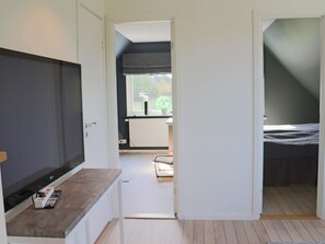 A room with a TV, a small table with a remote, a bed, and a window with a view of greenery.
