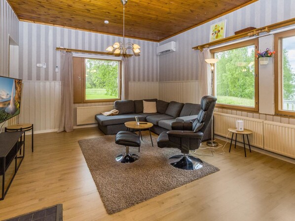 A modern living room with a sectional sofa, a coffee table, and a recliner. There are large windows providing natural light and a view of greenery outside.