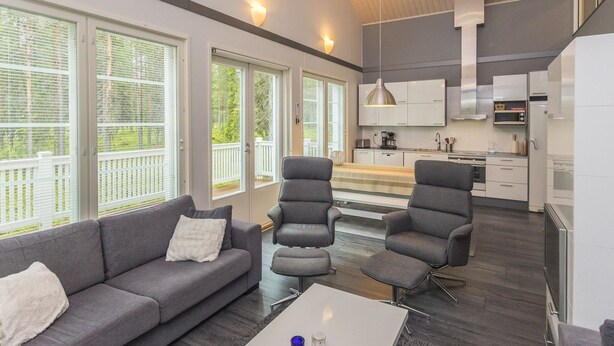Een moderne woonkamer met een grijze bank, twee fauteuils en een salontafel. Er zijn grote ramen met zonwering, een keuken met witte kasten en een houten plafond.