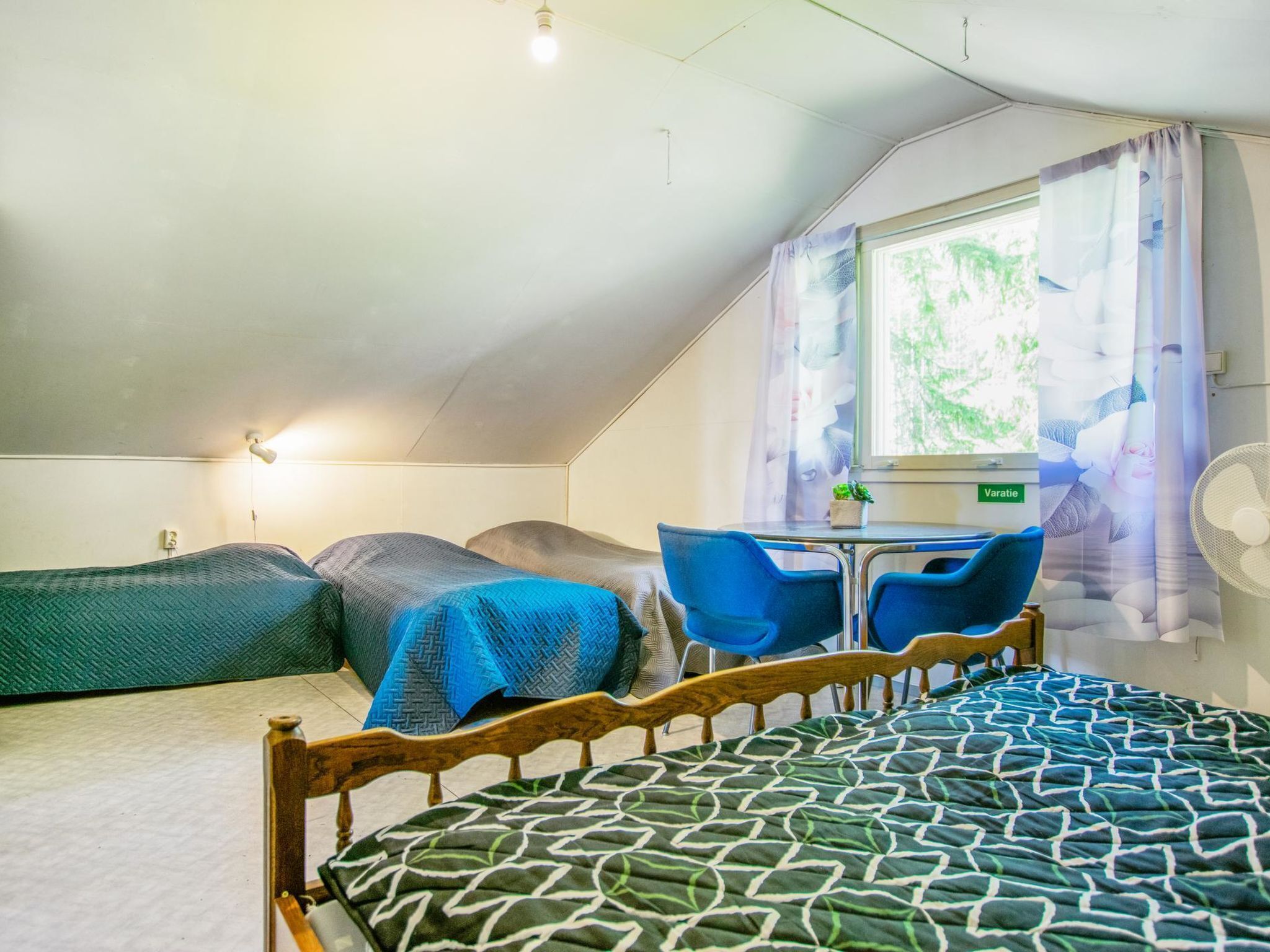 A small, cozy room with a bed, a desk, and two chairs. There is a window with curtains and a fan on the wall.