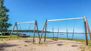 Ein sandiger Strand mit vier hölzernen Schaukeln mit Blick auf einen ruhigen See.