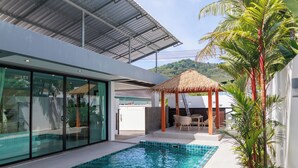 A modern outdoor pool area with glass doors, a thatched-roof gazebo, and a view of a lush hill.