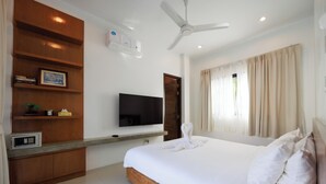A modern hotel room with a large bed, a flat-screen TV mounted on the wall, a ceiling fan, and a wooden shelf with books and a framed picture.