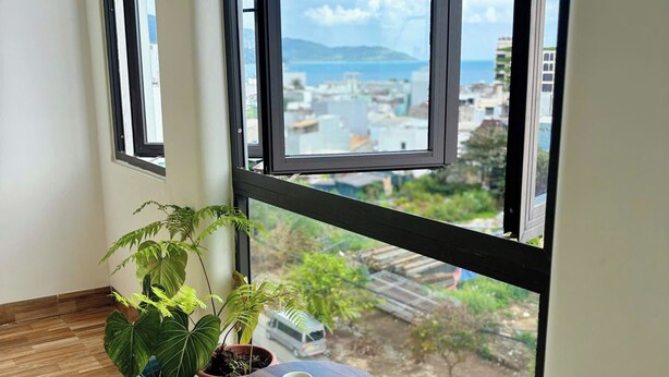 A room with a large window, a potted plant, a small table with a cup, and a sofa. - Ania Beachside Hotel Da Nang (Da Nang)