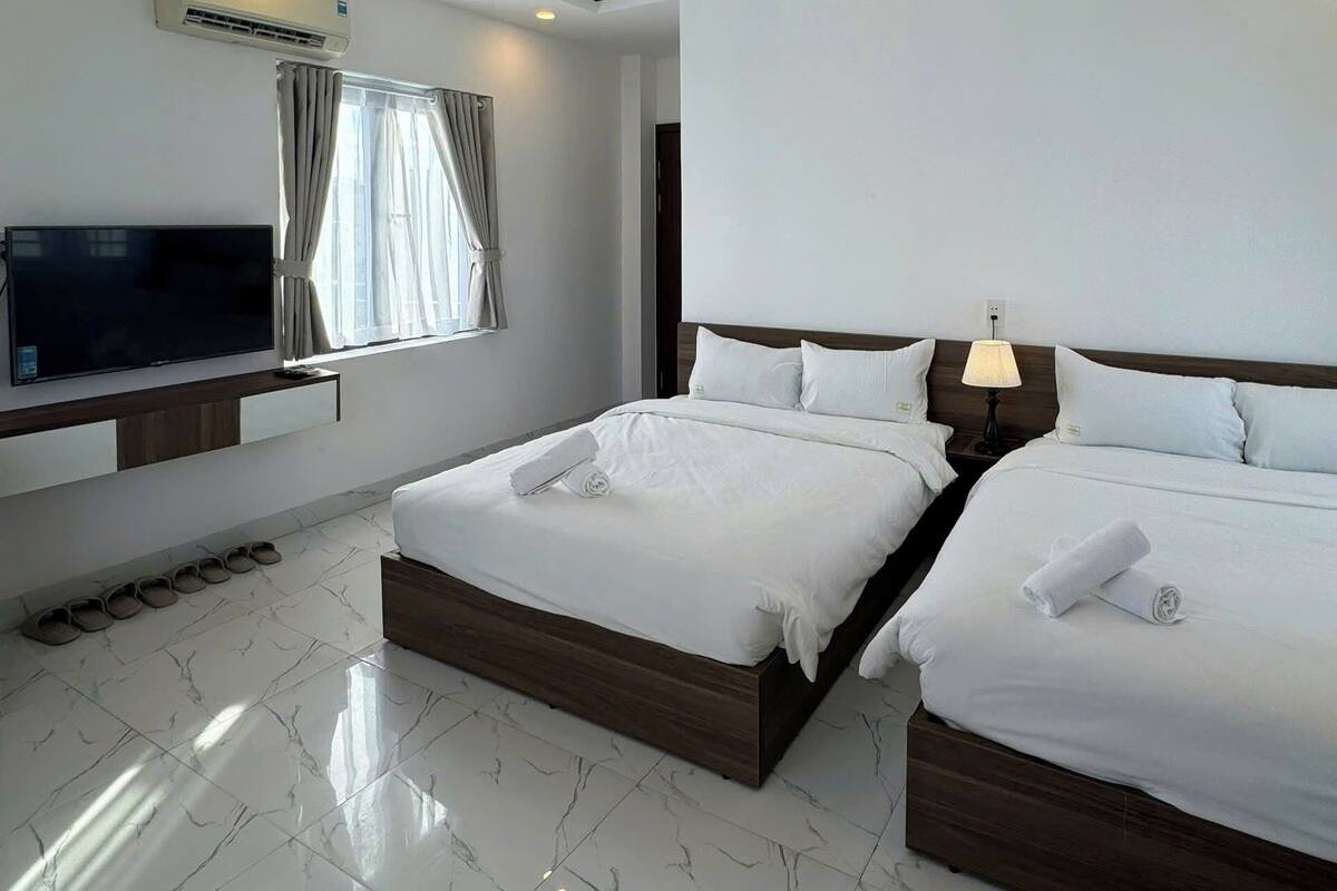 A hotel room with two beds, a flat-screen TV, an air conditioning unit, and a window with curtains.