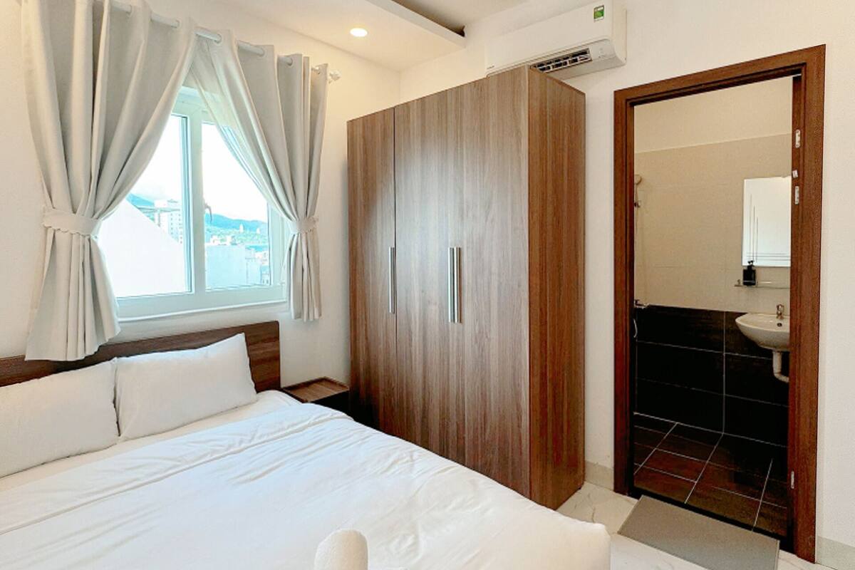 A hotel room with a large bed, a wardrobe, a bathroom with a sink, and a window with curtains.