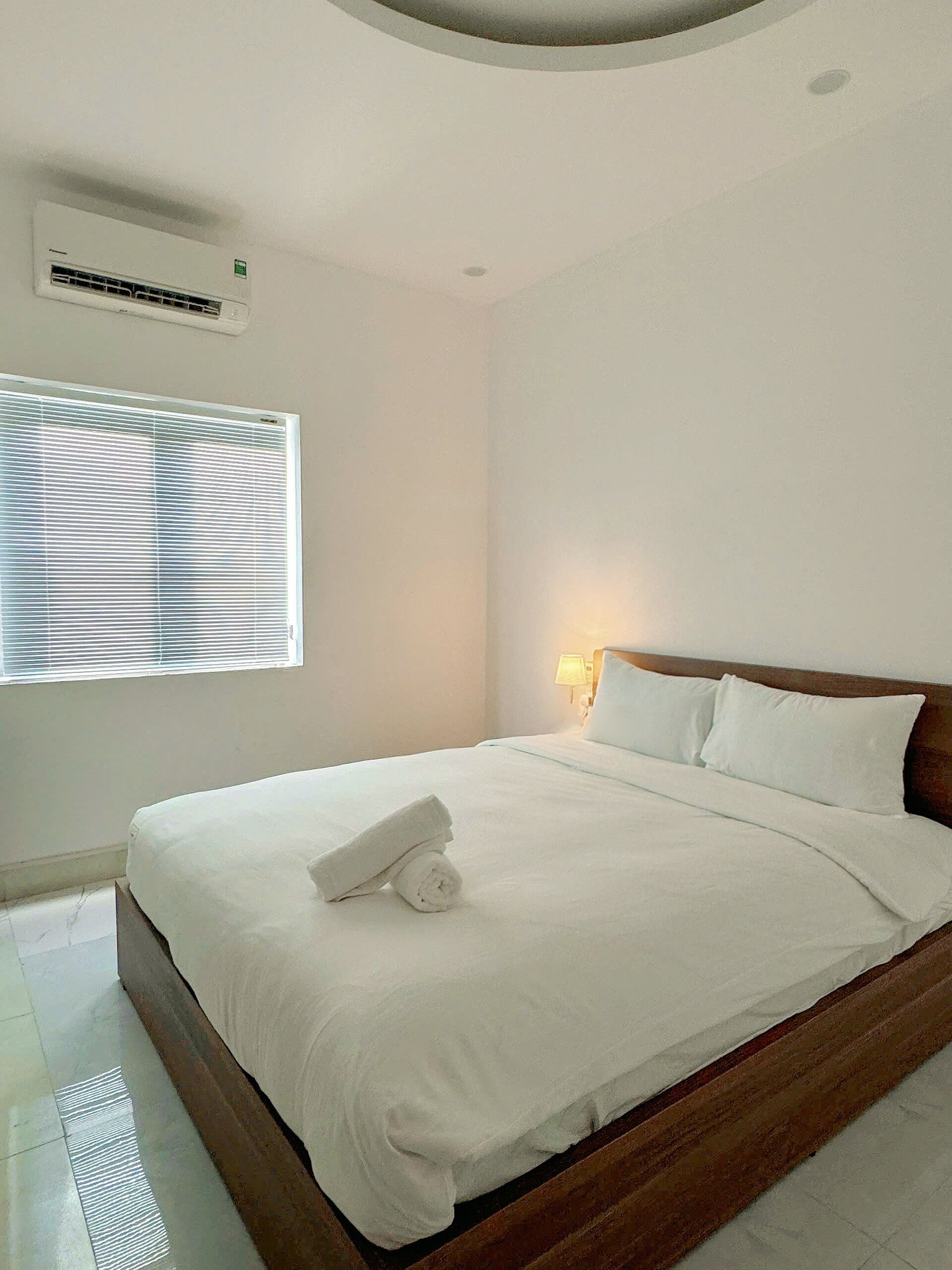 A hotel room with a large bed, a window with blinds, a wall-mounted air conditioner, and a bedside lamp.
