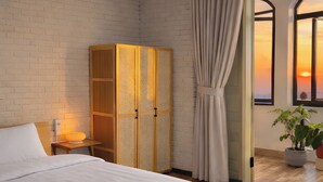 A bedroom with a wooden ceiling, a brick wall, a large bed with white bedding, a wooden wardrobe, and a view of the sunset through a window. - Ania Beachside Hotel Da Nang (Da Nang)