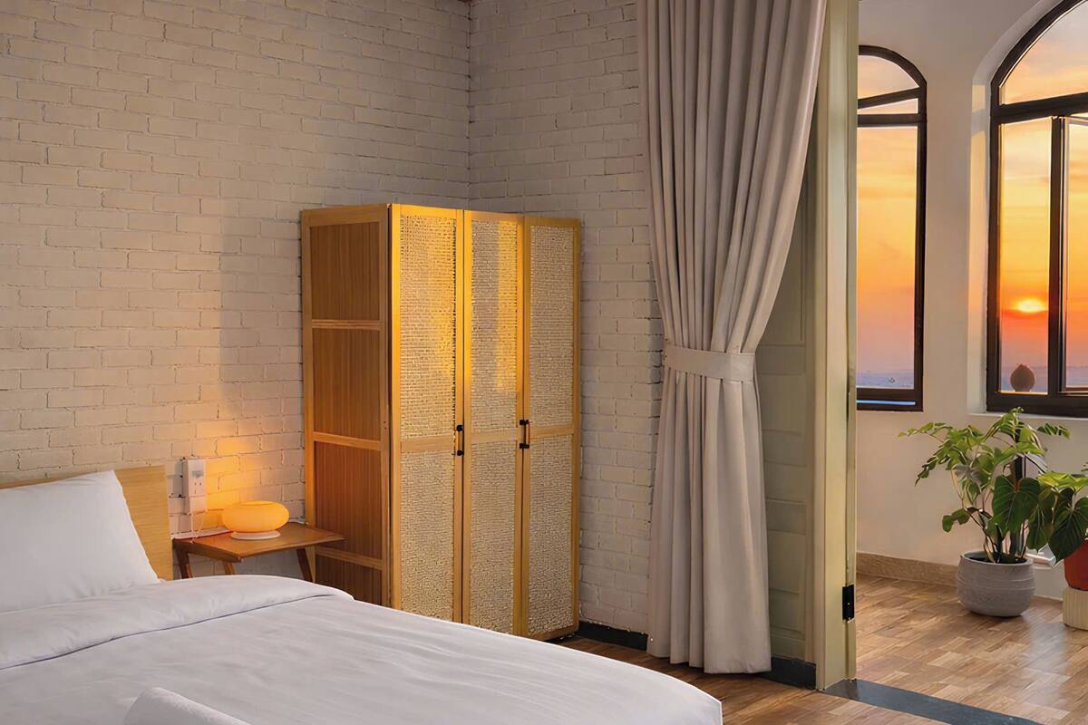 A bedroom with a wooden ceiling, a brick wall, a large bed with white bedding, a wooden wardrobe, and a view of the sunset through a window.