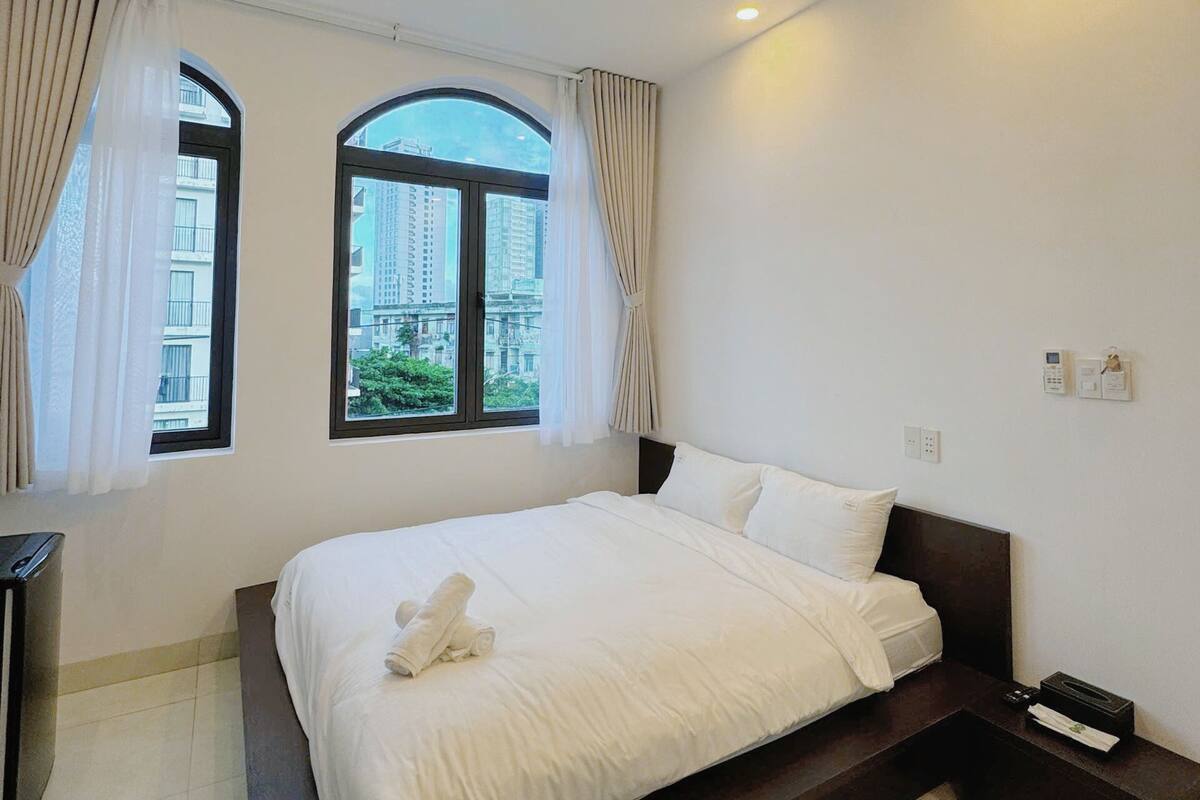 A hotel room with a large bed, two windows with curtains, a bedside table, and a view of buildings outside.